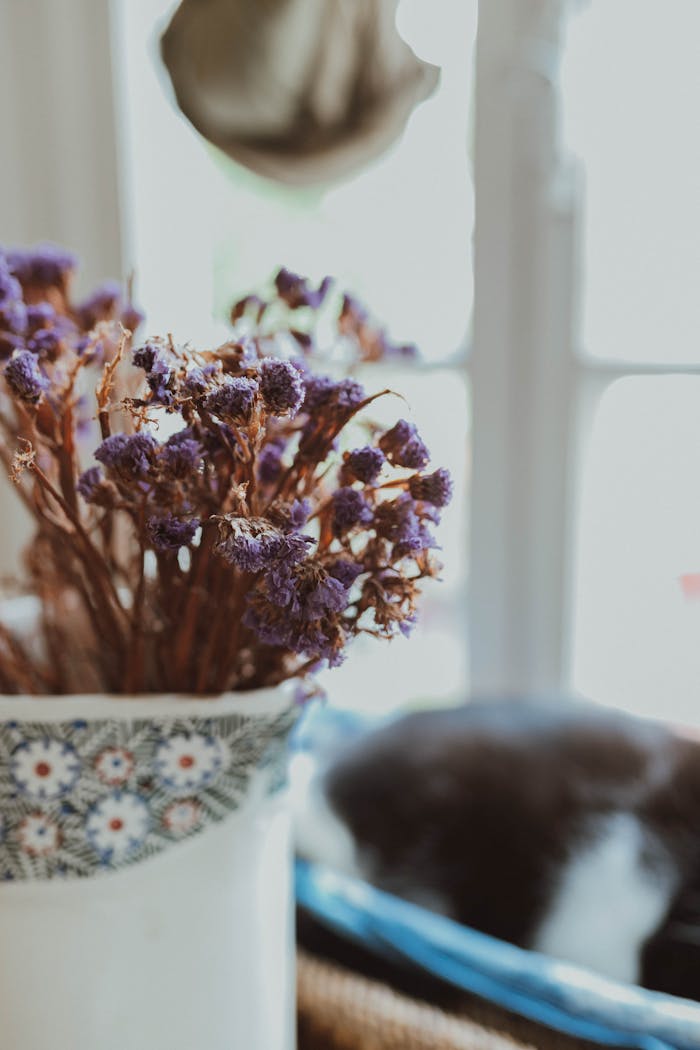 A vintage floral arrangement with dried purple flowers in a patterned vase by the window.
